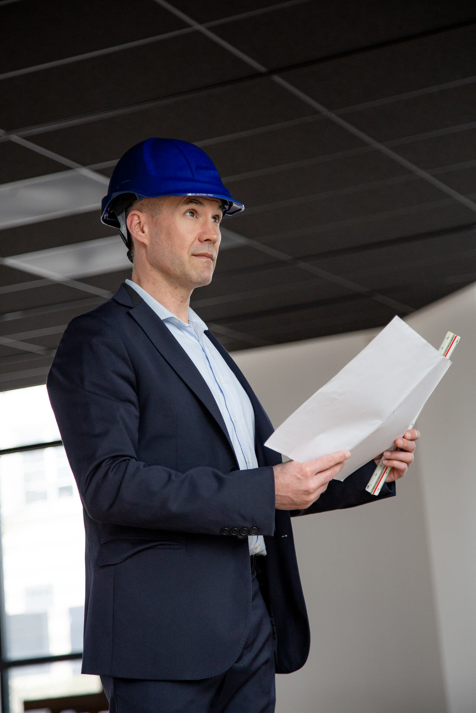 Nicolas DORGLER contractant général du bâtiment en Seine-et-Marne (77) en costume et casque de chantier, tenant un plan à la main et observant un chantier.